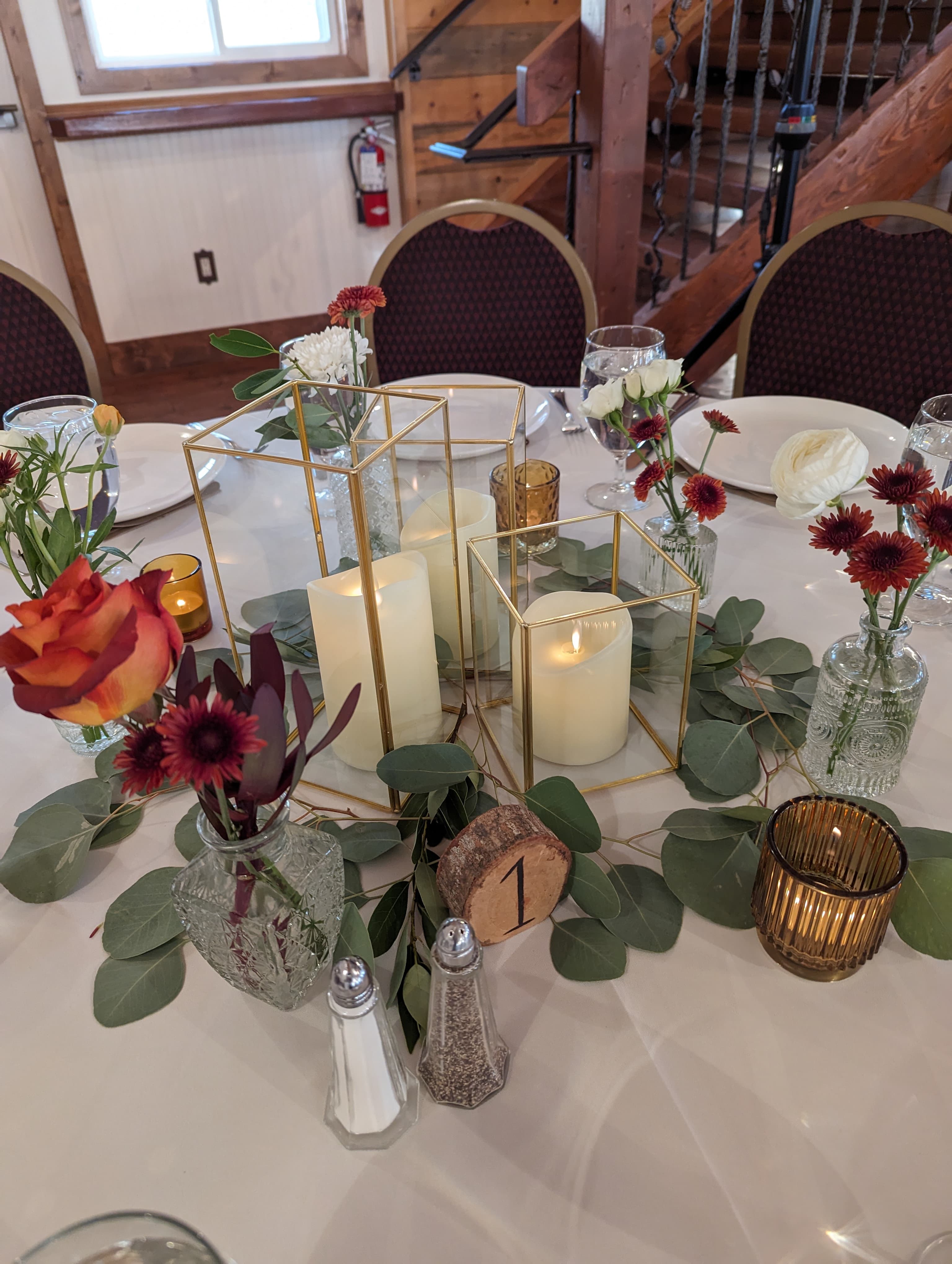 Reception table centerpiece with gold geometric terrarium candle holders, small bud vases with burgundy and orange blooms, and eucalyptus