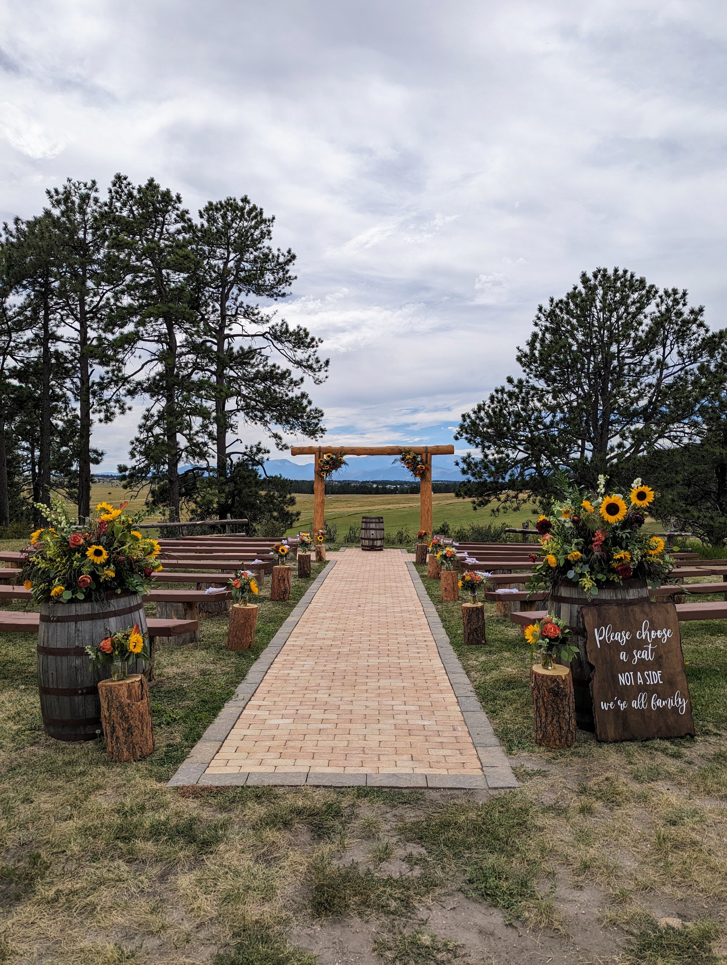 Outdoor ceremony setup with wooden benches, brick path aisle, wooden log arch with sunflowers, and mountain landscape in the background